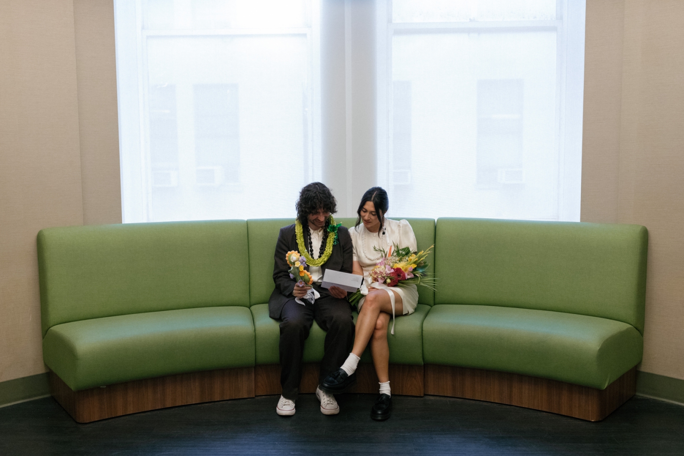 A bride and groom sit on a green couch at city hall, looking at their marriage license